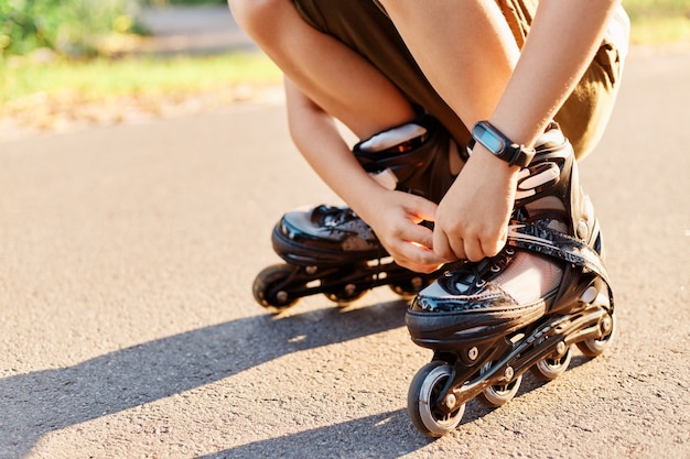 Portrait en gros plan d'un enfant inconnu s'accroupissant sur la route et fixant des lacets sur des patins à roulettes avant de patiner, un enfant sans visage s'amusant en plein air, faisant du patin à roues alignées, jouant seul.