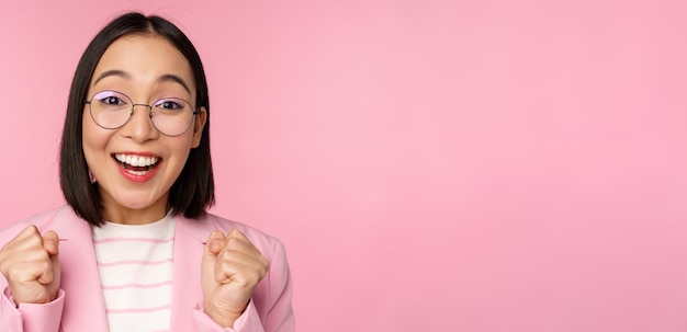 Portrait en gros plan d'une femme d'affaires asiatique acclamant l'enracinement pour regarder avec espoir et excitation à la caméra en souriant et en riant debout sur fond rose