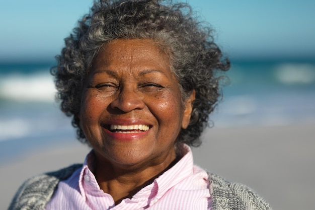 Portrait en gros plan d'une femme afro-américaine âgée debout sur la plage avec le ciel bleu et la mer en arrière-plan, souriant à la caméra