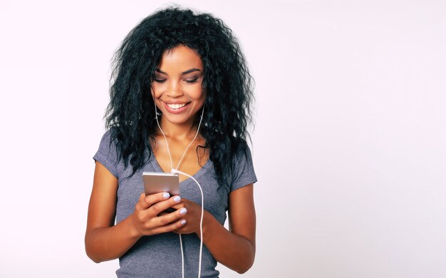 Portrait en gros plan d'une femme afro avec un large sourire séduisant et de longs cheveux crépus, qui exprime son bonheur en lisant quelque chose sur son smartphone