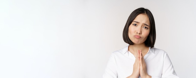 Portrait en gros plan d'une femme asiatique mendiant demandant smth dire s'il vous plaît avec un visage triste timide besoin d'aide debout sur fond blanc