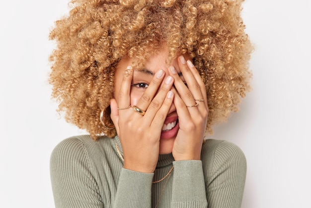 Portrait En Gros Plan D'une Femme Aux Cheveux Bouclés Heureuse Qui Regarde à Travers Les Doigts Cache Le Visage Exprime Des émotions Positives Vêtues De Vêtements Décontractés Isolés Sur Fond Blanc Se Sent Des Imbéciles Ludiques Autour