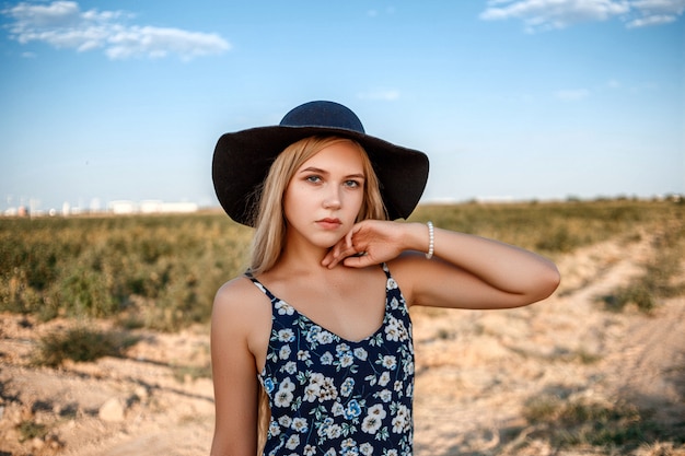 Portrait en gros plan d'une femme blonde aux yeux bleus portant un chapeau noir et une robe à imprimé floral. Elle pose dans un vignoble au coucher du soleil