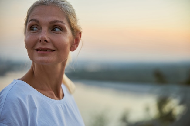 Portrait en gros plan d'une femme blonde mature et rêveuse souriante regardant quelque chose au loin