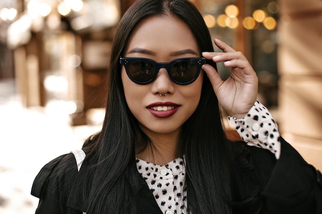 Portrait en gros plan d'une femme brune bronzée joyeuse et heureuse met des lunettes de soleil Dame asiatique en trench-coat noir sourit à l'extérieur