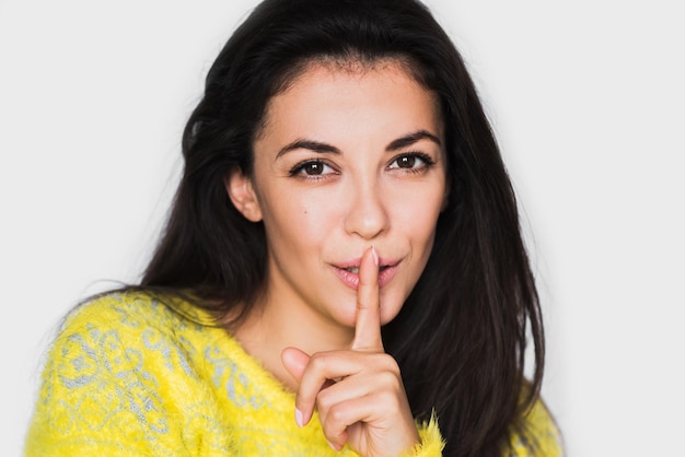 Portrait en gros plan d'une femme brune gaie montrant un geste silencieux avec le doigt sur la lèvre Jeune jolie femme heureuse portant un pull jaune sur fond gris clair Signe silencieux