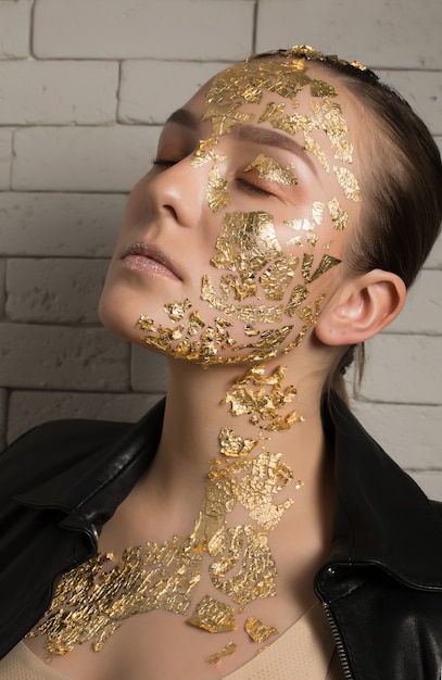 Portrait en gros plan d'une femme brune glamour avec une feuille d'or sur le visage et le cou, posant les yeux fermés