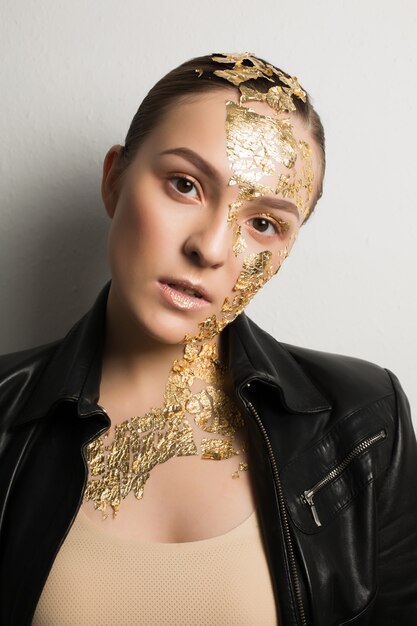 Portrait en gros plan d'une femme brune luxueuse avec une feuille d'or sur le visage et le cou, portant une veste en cuir