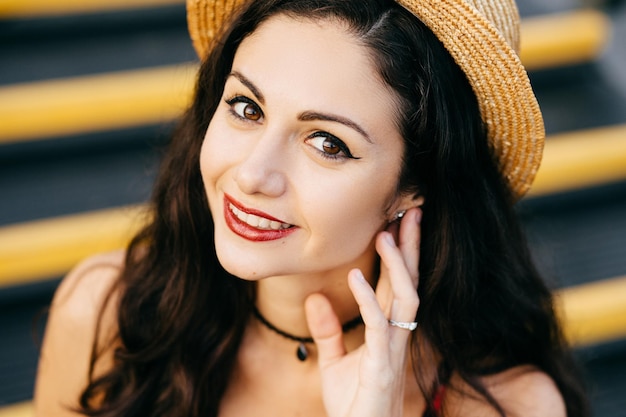 Portrait en gros plan d'une femme brune regardant avec ses yeux sombres lèvres peintes en rouge dans la caméra portant un chapeau de paille en gardant sa main sur l'oreille tout en étant assis dans les escaliers Belle femme avec un sourire charmant