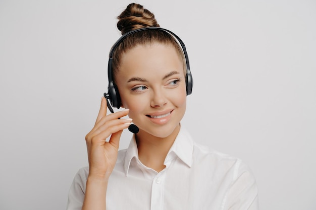 Portrait en gros plan d'une femme employée de banque souriante en chemise blanche et les cheveux en bouillon parlant sur un casque noir partageant des informations sur les clients regardant de côté isolé sur un fond gris