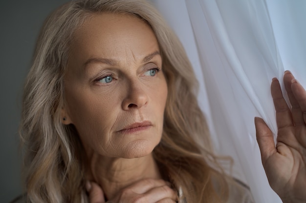 Portrait en gros plan d'une femme inquiète sérieuse touchant un rideau de fenêtre et regardant au loin