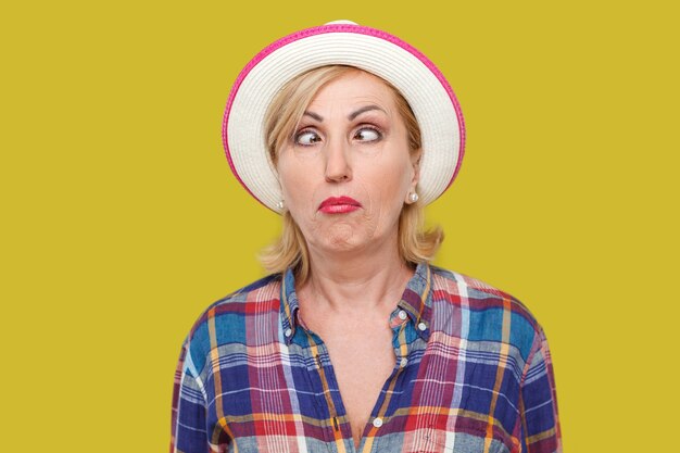 Portrait en gros plan d'une femme mature élégante et folle aux yeux croisés dans un style décontracté avec un chapeau blanc debout, les yeux croisés et regardant avec une expression de visage drôle. studio intérieur tourné isolé sur fond jaune.