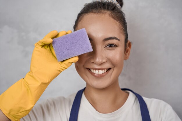 Portrait en gros plan d'une femme de ménage drôle dans des gants en caoutchouc jaune couvrant un œil avec une éponge de cuisine, regardant la caméra et souriant, debout contre un mur gris. Ménage, services de nettoyage