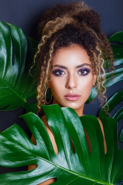 Photo portrait en gros plan d'une femme noire afro-américaine avec maquillage de soirée et feuilles tropicales sur mur noir