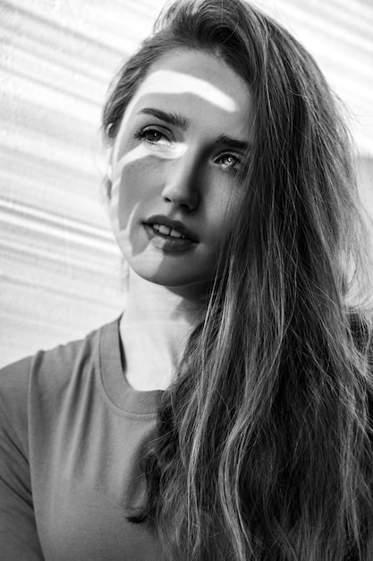 Portrait en gros plan d'une femme séduisante avec un maquillage naturel regardant loin avec une expression romantique et rêveuse. Photographie en noir et blanc, prise de vue en studio intérieure éclairée par la lumière du soleil depuis la fenêtre.