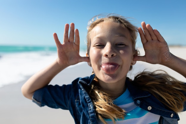 Portrait en gros plan d'une fille caucasienne sur une plage ensoleillée, faisant une grimace drôle et souriant à la caméra, avec le ciel bleu et la mer en arrière-plan