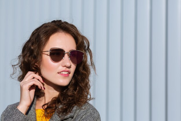 Portrait en gros plan d'une fille hipster élégante portant des lunettes de soleil et un manteau, posant près des volets. Espace pour le texte