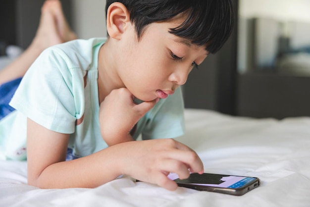 Portrait en gros plan d'un garçon asiatique avec une frange noire, des yeux noirs, un visage souriant, portant une chemise vert clair, allongé sur le lit de sa maison, jouant avec son téléphone intelligent.