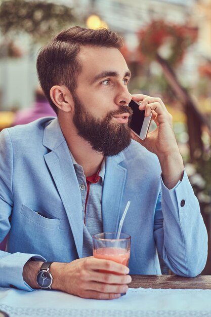 Portrait en gros plan d'un homme d'affaires barbu à la mode avec une coupe de cheveux élégante, parlant par téléphone, boit un verre de jus frais, assis dans un café à l'extérieur.