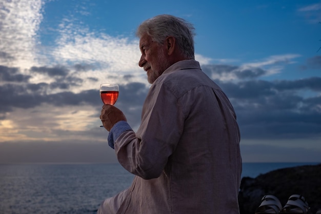 Portrait en gros plan d'un homme âgé assis devant la mer au coucher du soleil tenant un verre de vin horizon au-dessus de l'eau Sentiment positif pour les hommes âgés profitant de vacances ou de retraite