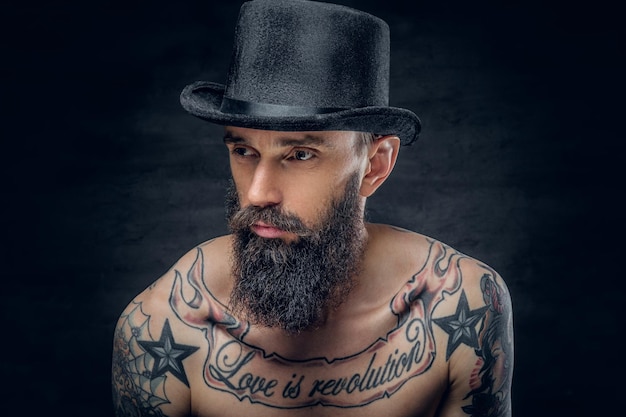 Portrait en gros plan d'un homme barbu en chapeau haut de forme avec des tatouages sur sa poitrine sur fond sombre.