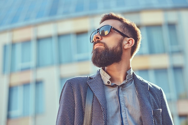 Portrait en gros plan d'un homme barbu portant des vêtements décontractés et des lunettes de soleil, debout dans une rue de la ville contre un gratte-ciel.