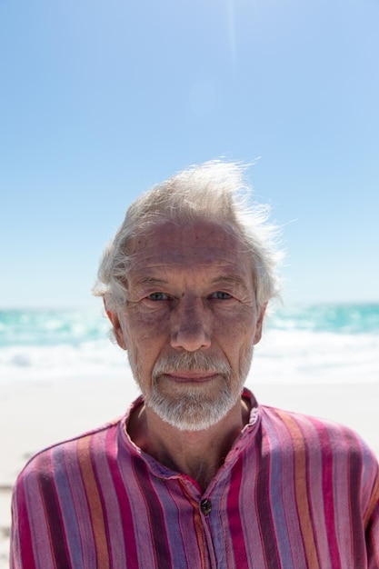 Portrait en gros plan d'un homme caucasien âgé debout sur la plage avec le ciel bleu et la mer en arrière-plan, regardant la caméra