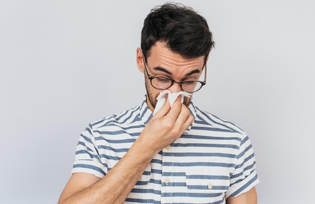 Portrait en gros plan d'un homme malade de race blanche portant une chemise rayée et des lunettes pris froid et éternuant dans les tissus L'homme a le virus des maux de tête sur fond blanc Concept de médecine saine