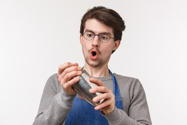 Portrait de gros plan d'un jeune employé dévoué et enthousiaste, le barman aime faire des boissons, secouant un cocktail dans un shaker avec passion, debout sur un mur blanc excité