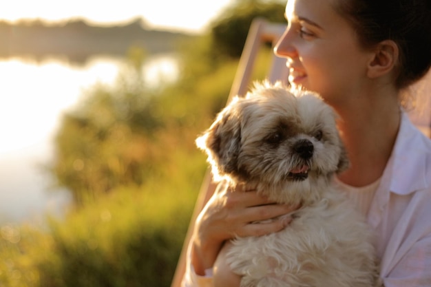 Portrait en gros plan d'une jeune femme adulte séduisante portant une chemise blanche assise avec son chien pékinois près de la rivière et regardant ailleurs avec un sourire charmant se reposant en profitant de la belle nature et du coucher du soleil