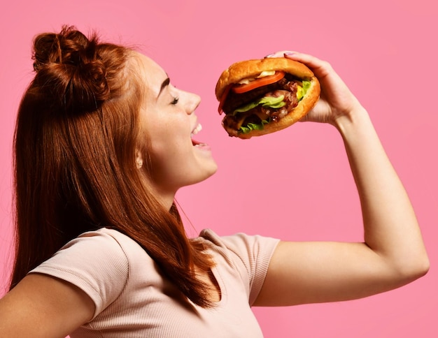 Portrait en gros plan d'une jeune femme affamée mangeant un hamburger isolé sur fond rose
