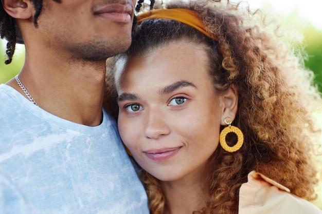 Portrait en gros plan d'une jeune femme aux cheveux bouclés souriant à la caméra et embrassant un petit ami anonyme duri