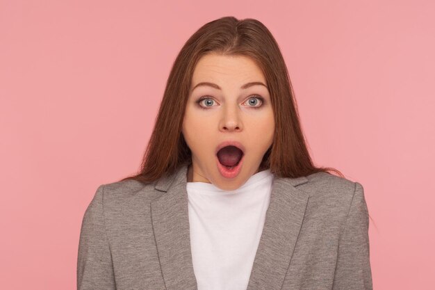 Portrait en gros plan d'une jeune femme brune étonnée en costume d'affaires regardant la caméra avec la bouche ouverte regardant avec surprise exprimant un véritable étonnement incrédulité studio intérieur tourné sur fond rose