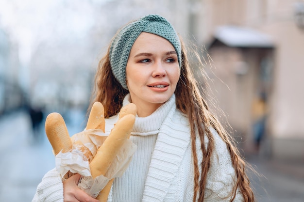 Portrait en gros plan d'une jeune femme en costume en tricot blanc tenant des petits pains. Expression langoureuse sur son visage et détourner le regard. Contexte de la ville de bokeh. Boulangeries publicitaires et un espace ouvert pour le texte.