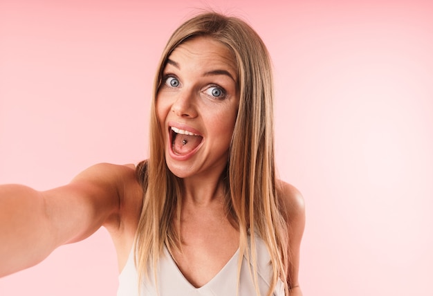 Portrait en gros plan d'une jeune femme étonnée souriant à la caméra tout en prenant un selfie Portrait isolé sur un mur rose