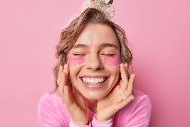 Portrait en gros plan d'une jeune femme joyeuse aux cheveux ondulés touche le visage garde doucement les yeux fermés sourit largement subit des procédures de beauté applique des patchs d'hydrogel sous les yeux veut être belle