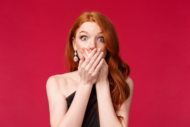 Portrait en gros plan d'une jeune femme rousse sans voix, choquée et inquiète a découvert le terrible secret de quelqu'un, les commérages, le souffle coupé et la bouche avec les mains, debout sur un mur rouge