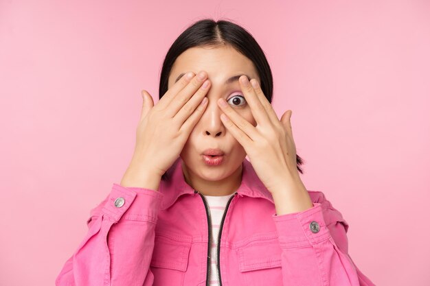 Portrait en gros plan d'une jeune fille asiatique qui a l'air surprise d'exprimer son étonnement et son émerveillement en regardant à travers les doigts debout sur fond rose