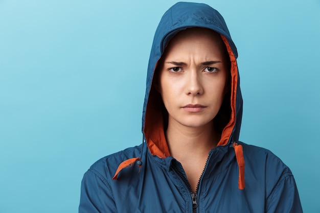 Portrait en gros plan d'une jeune fille contrariée portant une capuche, isolée sur un mur bleu, regardant la caméra