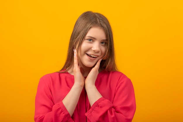 Portrait en gros plan d'une jeune fille souriante gentiment et faisant les yeux jeune femme en chemise rouge sur fond jaune