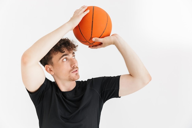 Portrait en gros plan d'un jeune homme concentré en t-shirt tenant et lançant une balle tout en jouant au basket-ball isolé sur un mur blanc