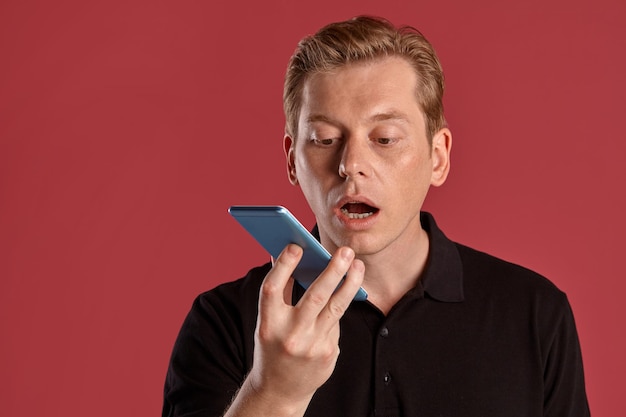 Portrait en gros plan d'un jeune homme élégant au gingembre dans un t-shirt noir élégant parlant par un smartphone tout en posant sur fond de studio rose. Expressions faciales humaines. Concept d'émotions sincères. Copier l'espace