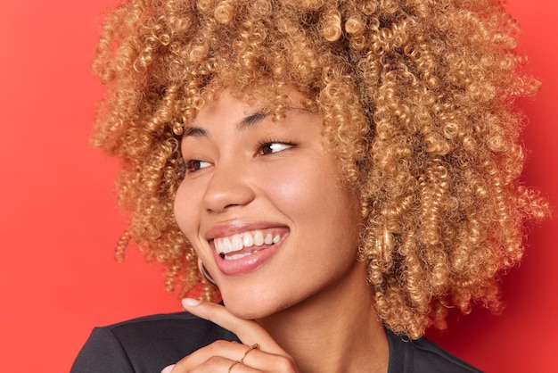 Portrait en gros plan d'une jeune jolie femme positive avec une peau saine sans maquillage et des cheveux touffus bouclés touchent le menton doucement concentré loin heureusement isolé sur fond rouge. Notion de bonheur