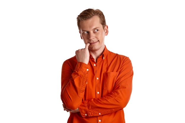 Portrait en gros plan d'un jeune mec séduisant au gingembre dans une chemise orange élégante agissant comme complotant quelque chose tout en posant isolé sur fond de studio blanc. Expressions faciales humaines. Émotions sincères