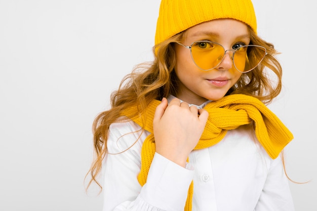 Portrait en gros plan d'une jolie adolescente blonde souriante dans un chapeau jaune et des lunettes de soleil, dans une chemise blanche avec un pull noué sur ses épaules sur un blanc avec copie espace