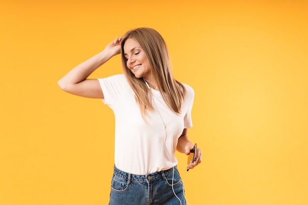 Portrait en gros plan d'une jolie femme blonde souriante tout en écoutant de la musique avec un smartphone et des écouteurs isolés sur un mur jaune