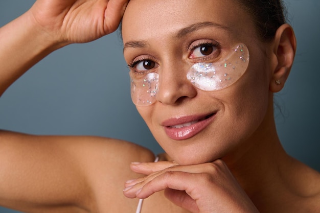 Portrait en gros plan d'une jolie femme souriante appliquant des patchs oculaires en collagène d'hydrogel médical doré pour réduire les poches sous les yeux et les poches, posant à la caméra sur un fond gris. Copier l'espace