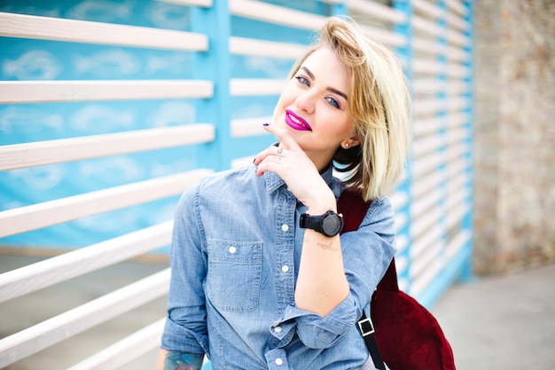 Portrait en gros plan d'une jolie fille blonde aux lèvres rose vif et aux yeux bleus portant une chemise en jean bleu et un sac à dos marsala avec des rayures bleues et blanches en arrière-plan.