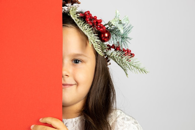 Portrait en gros plan d'une jolie fille dans une couronne de Noël cachant la moitié du visage derrière une bannière vierge rouge isolat...