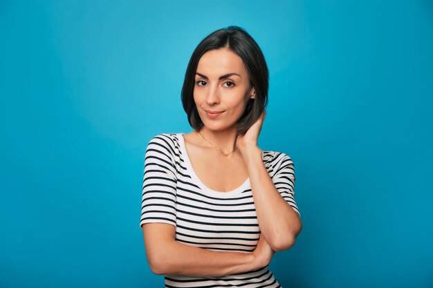 Portrait en gros plan d'une jolie jeune femme brune souriante alors qu'elle pose dans un t-shirt rayé avec les bras croisés et regardant la caméra isolée sur fond bleu